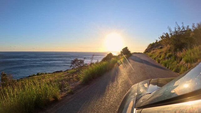 LENS FLARE: Evening sun hangs low over horizon, casting golden light onto coastal road that winds above blue sea. View from side of the car offers a unique perspective on morning journey around island