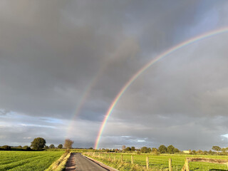 A stunning and colorful double rainbow beautifully arches over lush green fields that lie beneath a cloudy sky