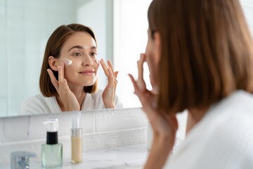 Beautiful woman applying moisturizing cream to her face for daily skincare routine in bright bathroom