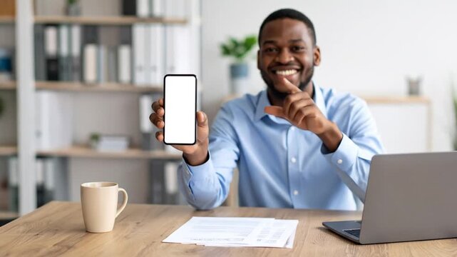Smiling African American Man Holding Smartphone with White Screen Displayed in Bright Office with Laptop Mug and File Folders