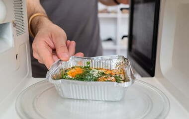 Person placing ready-to-eat meal in aluminum tray into a microwave oven for heating.