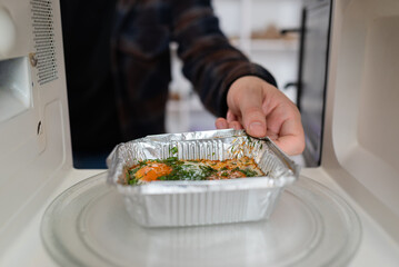 A person's hand places an aluminum takeaway container with food into a microwave, a common kitchen...