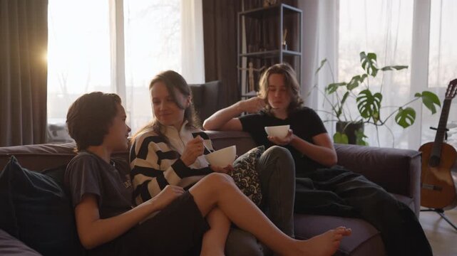A happy family with a mother and two teenage sons sits together on a sofa at home, eating a snack from bowls. They communicate, laugh, and relax, enjoying quality time and bonding in the living room.