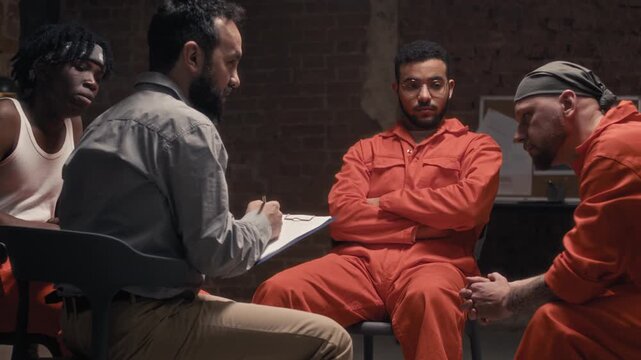 Medium shot of mature male prison psychologist talking to group of diverse attentive convicts during support therapy session, giving advice on processing negative emotions and experience