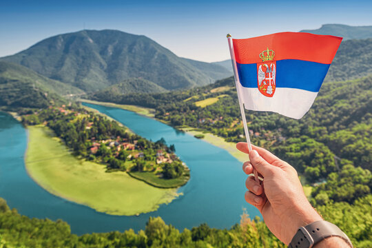 Hand waving Serbian flag with scenic view of meandering river and green islet with houses and mountains in background