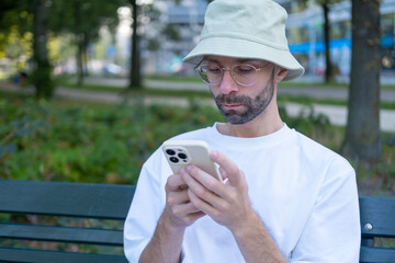 Fototapeta premium Young man checking smartphone on park bench in city, Millennial guy sitting outside texting or reading news, Modern lifestyle concept man connected online on go, quiet moment, modern city life