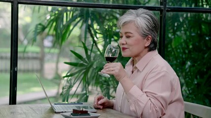 asian senior female reacting with cheerful expression raising fists smiling joyfully while reading something on laptop during solo cafe moment with wine and dessert after travel meetup