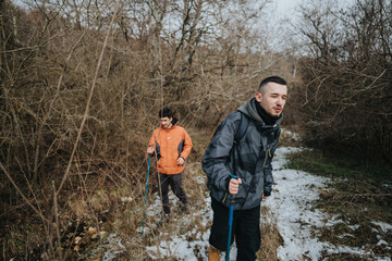 Two hikers traverse the snowy forest trail, surrounded by bare trees in cold winter weather. Their...
