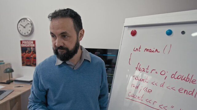 Medium close-up of mature Caucasian male teacher writing lines of computer code on white board and explaining programming rules while conducting lesson in penitentiary institution