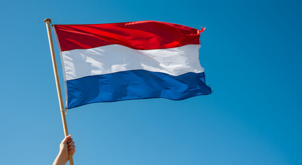 Hand holding a waving red, white, and blue flag against a vibrant blue sky