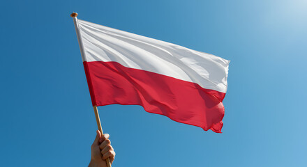 Hand holding a white and red national flag waving in the wind against a clear blue sky, symbolizing national pride.