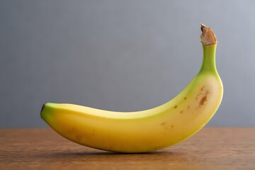 A single ripe yellow banana with brown spots on a wooden surface with a grey background