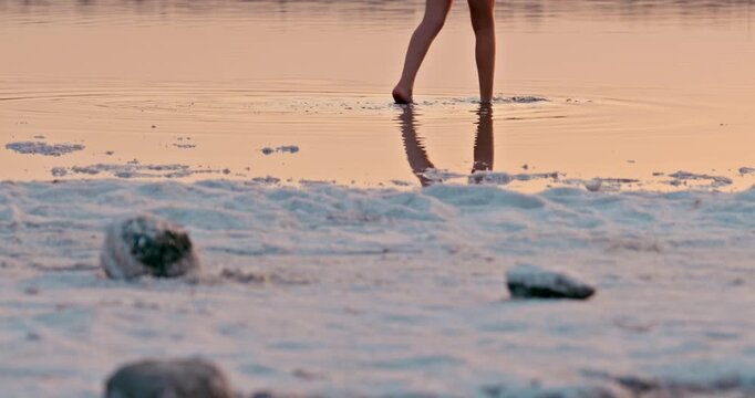 A little girl walks barefoot along a salt-covered shore beside her mother during sunset. A warm, intimate moment of connection in a serene natural setting.