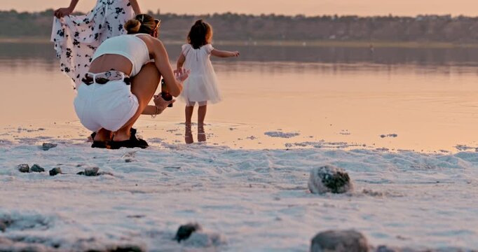 A little girl walks barefoot along a salt-covered shore beside her mother during sunset. A warm, intimate moment of connection in a serene natural setting.