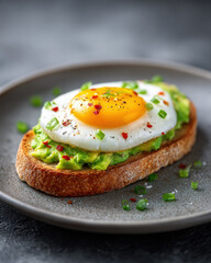 Avocado toast with fried egg on whole grain bread, garnished with chives and chili flakes, served on gray plate