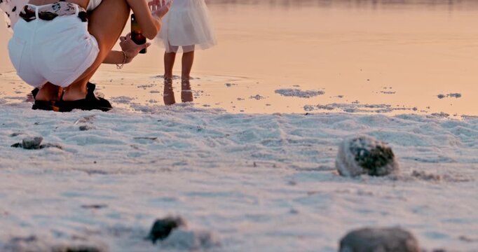 A little girl walks barefoot along a salt-covered shore beside her mother during sunset. A warm, intimate moment of connection in a serene natural setting.