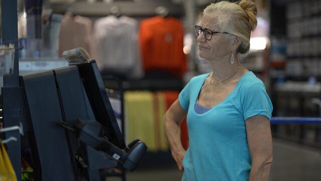 In a hardware store, a frustrated confused mature woman is using a self checkout station, scanning items with a handheld scanner while preparing to complete her purchase.