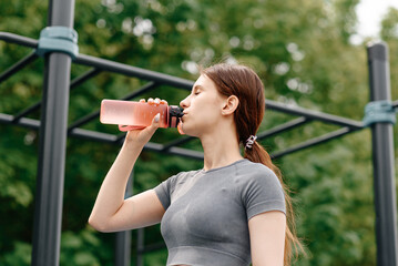 young woman stands on street sports ground before doing sport exercises outdoors in summer, drinks...