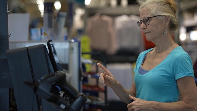 A mature woman engages with a self checkout machine in a busy hardware store, using her credit card trying to figure out how to finalize her shopping transaction.
