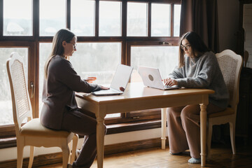 Two adult student study on-line on laptops at a table near a large window, discussion or working side-by-side. Comfortable, domestic atmosphere, remote education, or webinar