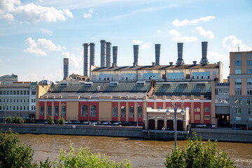 An old building of a former red brick thermal power plant with pipes on the embankment of the Clear sunny day river, Moscow.  Sights of Russia. Architecture of World tourism.