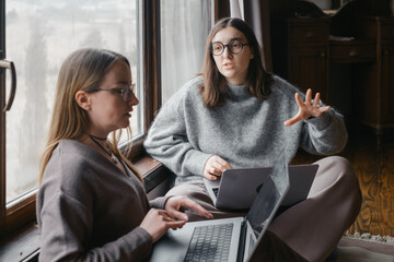 Two women engaged in a discussion while sitting indoors with laptops. Collaborating on a project, sharing ideas in comfortable setting. Work, study or team environments.