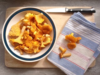 Top view of a porcelain bowl full of chanterelles. The bowl is on a wooden cutting board with a kitchen towel, a knife and a few more mushrooms.