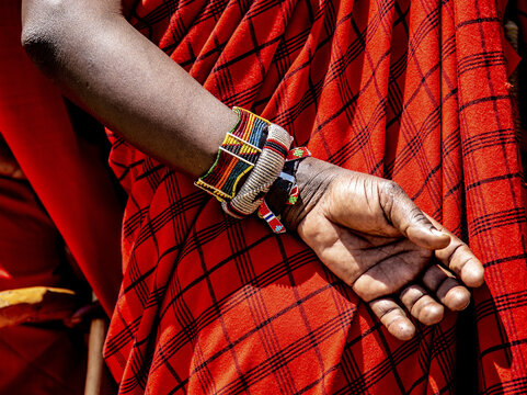 View of a person's arm adorned with vibrant beaded bracelets against a striking red checkered fabric, rich in texture and cultural significance, Narok, Narok, Kenya.