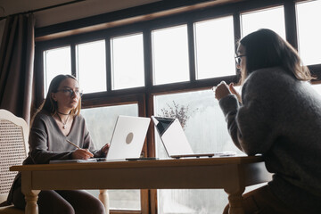 Two adult student study on-line on laptops at a table near a large window, discussion or working side-by-side. Comfortable, domestic atmosphere, remote education, or webinar