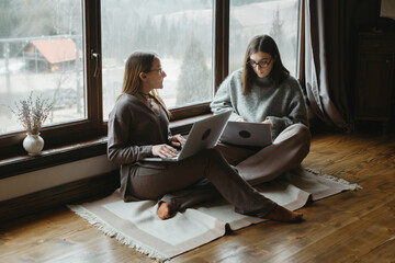 Two women freelancers are sitting near large window, working on their laptop. Friends collaborate or share ideas. Small team communication, work remotely at home workspace.