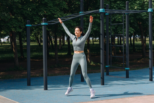 young slim caucasian woman doing jumping jack exercises outdoors in summer, street sports ground, grey sportswear, green trees in background, healthy lifestyle concept - Powered by Adobe