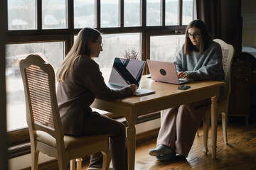Two adult student study on-line on laptops at a table near a large window, discussion or working side-by-side. Comfortable, domestic atmosphere, remote education, or webinar