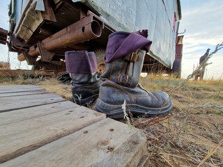 Worn-out work boots with rolled-up pants lie on dry grass near an old metal trailer. Rustic...