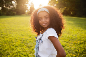 A cheerful young girl standing in a sunlit park, radiating joy and positivity. The scene captures a...