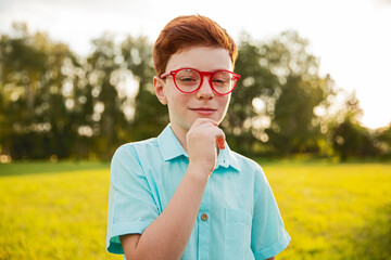 A young boy wearing red glasses and a blue shirt stands in a green park during a sunny day. The...