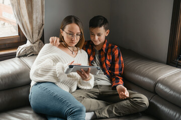 Teenager with mother playing on console at home. Woman and son sitting close together on leather couch in a relaxed, indoor setting.