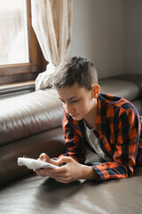 A boy is lying on a couch, absorbed in playing a video game. He is holding a white controller and appears focused on the screen. Natural light comes through a window with a curtain in the background.