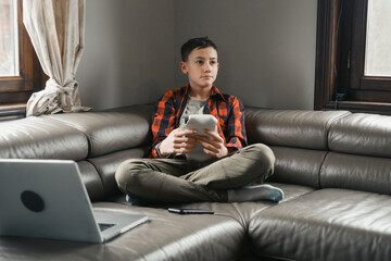 Teenager sitting with game console on the sofa. Boy playing video game instead of study. Digital detox and social media, addiction. Boy spending free time with gadgets