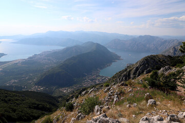 Stunning panoramic view of the Bay of Kotor in Montenegro, seen from a high mountain viewpoint. The landscape features deep blue waters, green hills, rugged rocky slopes, and scattered villages