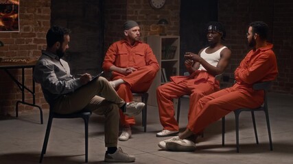 Full shot of young African American male inmate in orange uniform and white vest contemplating on criminal life at group therapy, while psychologist and multiethnic companions are listening