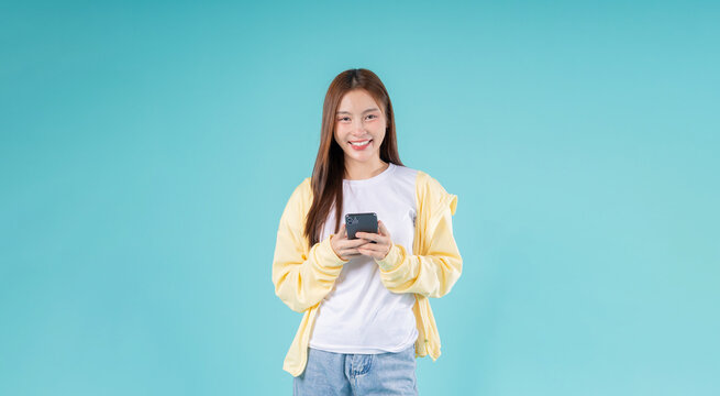 Happy young Asian woman using a smartphone while standing against a sky blue background, symbolizing modern communication, social media, digital lifestyle, and technology.