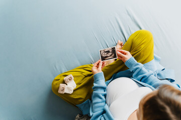 Pregnant woman holding ultrasound image in front of her baby bump, sitting on bed. Mother-to-be