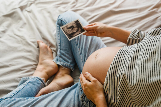 Pregnant woman holding ultrasound image in front of her baby bump, sitting on bed. Mother-to-be