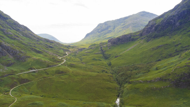 Aerial view of a winding road snaking through the vibrant green valley, cradled by majestic mountains under a soft sky, Glencoe, Scotland, United Kingdom.