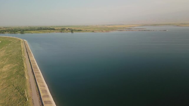 A dam wall runs beside calm blue water at Djizak Reservoir. Sunlight falls across green grass and the long straight edge. Distant countryside and fields stretch far under a clear, quiet sky