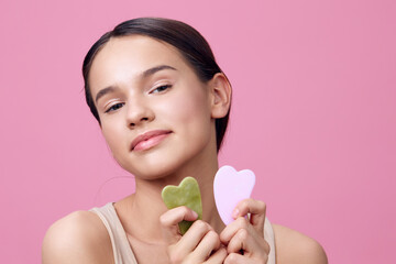 Young woman holding jade and rose quartz facial massagers, posing with a soft smile against a pink...