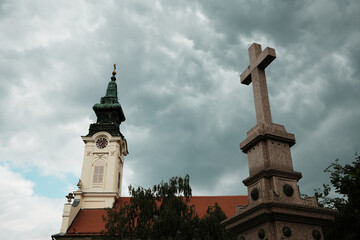Church tower and stone cross under stormy skies in Sombor, Serbia. A striking mix of history,...