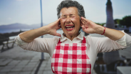 Senior woman with grey hair in apron covering ears on a seaside promenade with a worried...