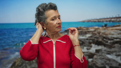Senior woman with grey hair in a vibrant red outfit touches her hair at a seaside location against...