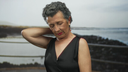 Senior woman with grey hair touching her neck in discomfort on a seaside promenade with a serene beach background in the distance.